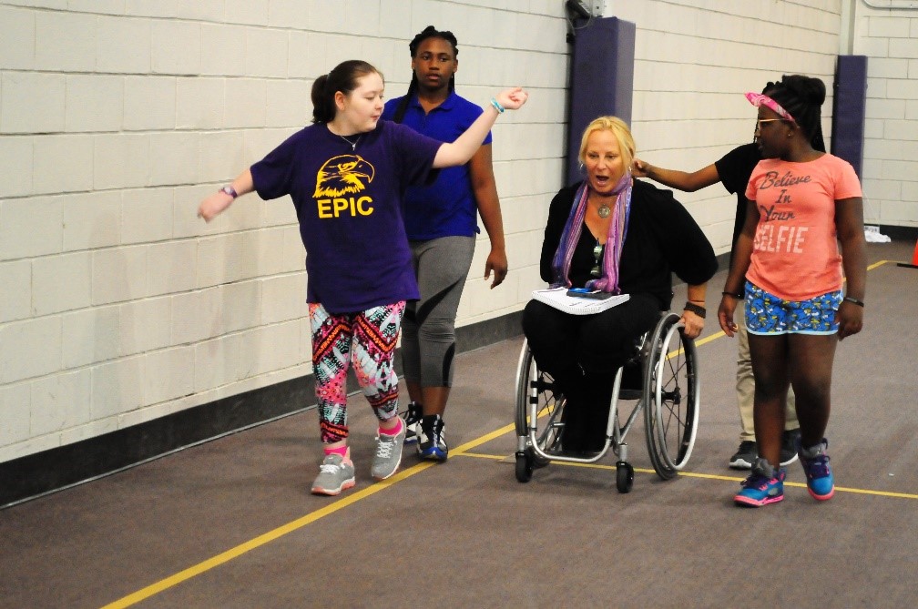 Four girls in running clothes and an adult in a wheelchair take laps around a gymnasium. 