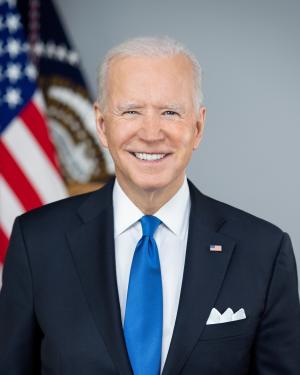 official photo of President Biden smiling and wearing a dark suit and a blue tie