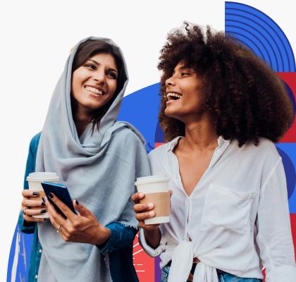Two women walking together and smiling while holding coffee. One woman wears a headscarf while the other woman has brown tight curly hair.