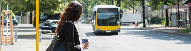 A woman wearing a business suit and holding a cup of coffee stands at a bus stop and watches a bus approach.